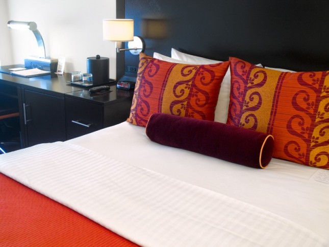 A hotel room bed with white sheets, orange and red pillows, and a black desk with a lamp.